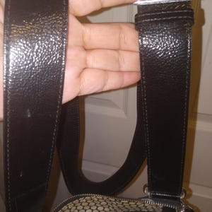 May include: Black leather shoulder bag with a textured finish. The bag features a silver-tone buckle and a patterned interior lining. The strap is wide and adjustable. The bag is held by a hand.