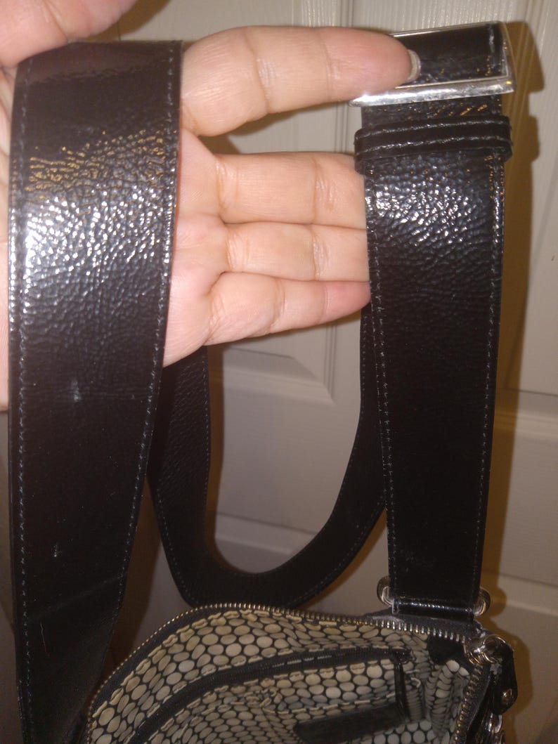 May include: Black leather shoulder bag with a textured finish. The bag features a silver-tone buckle and a patterned interior lining. The strap is wide and adjustable. The bag is held by a hand.