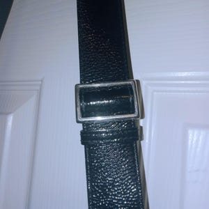 May include: Close-up of a black leather strap with a silver-colored rectangular buckle. The strap has a textured surface and appears to be part of a bag or accessory. The buckle is shiny and reflects light.