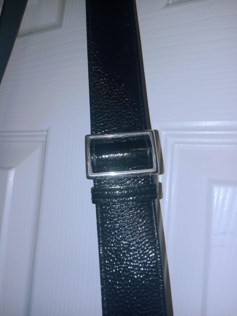 May include: Close-up of a black leather strap with a silver-colored rectangular buckle. The strap has a textured surface and appears to be part of a bag or accessory. The buckle is shiny and reflects light.