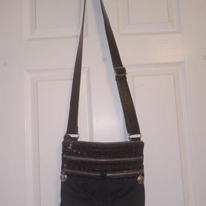 May include: Black crossbody bag with three zippered compartments and a long adjustable strap. The top section has a faux crocodile skin texture. Silver-toned hardware accents the bag, which is hanging on a white door.