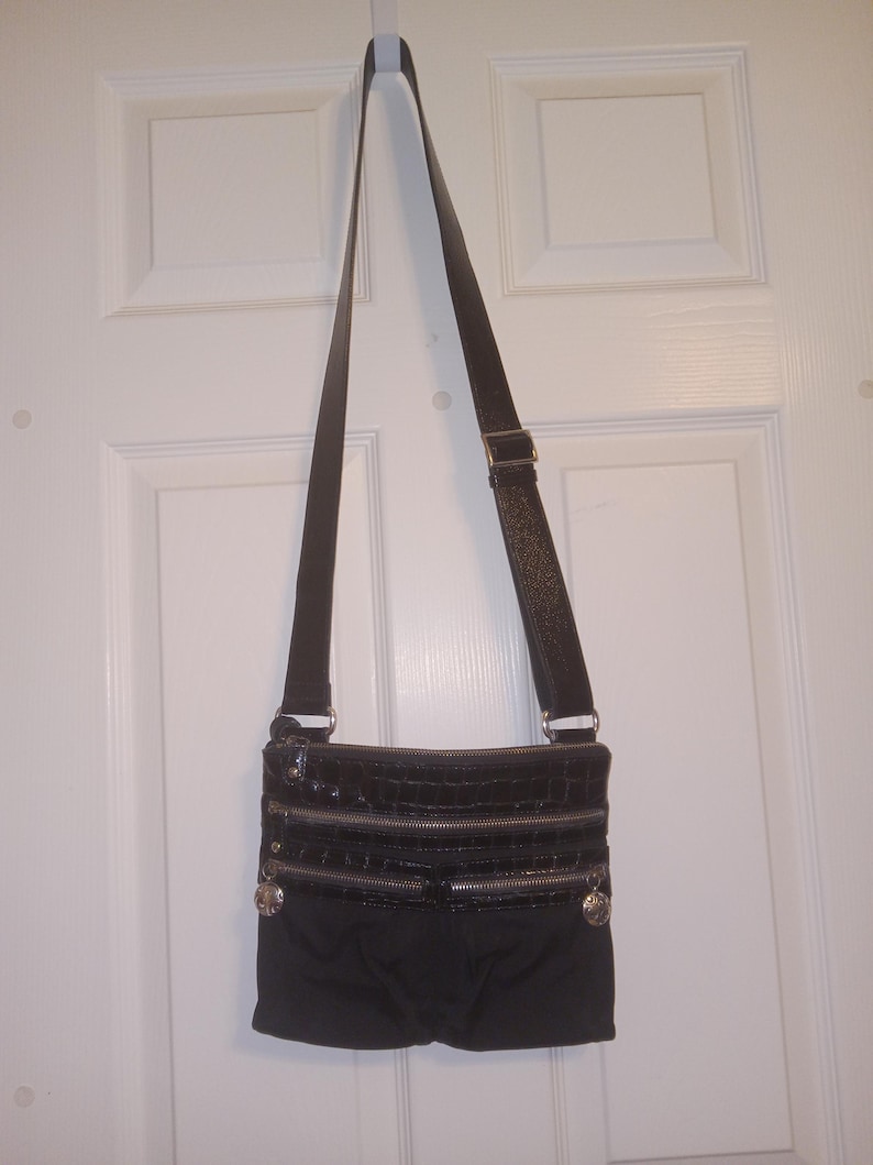 May include: Black crossbody bag with three zippered compartments and a long adjustable strap. The top section has a faux crocodile skin texture. Silver-toned hardware accents the bag, which is hanging on a white door.