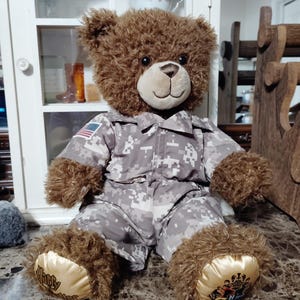 Könnte beinhalten: Ein brauner Plüschteddybär in einem grauen und weißen, digital gemusterten Camouflage-Militäranzug. Der Bär hat einen kleinen Aufnäher der amerikanischen Flagge am rechten Ärmel und goldfarbenen Stoff an den Fußsohlen mit Text und Wappen.
