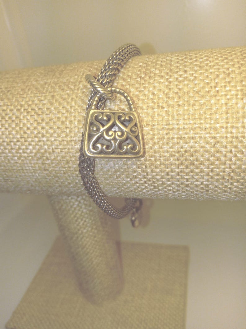 Pu&ograve; includere: Un braccialetto color argento con un ciondolo a forma di lucchetto decorativo. Il ciondolo presenta un design ornato a volute. Il braccialetto &egrave; esposto su una superficie beige strutturata, che mette in risalto i dettagli e la maestria del gioiello.