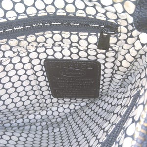 May include: Interior view of a black handbag with a white and black polka dot lining. A black zipper and a black leather tag with embossed text are visible. The bag's interior is spacious and well-organized.