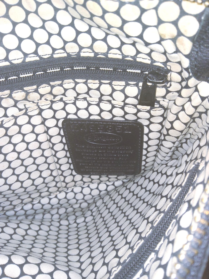 May include: Interior view of a black handbag with a white and black polka dot lining. A black zipper and a black leather tag with embossed text are visible. The bag's interior is spacious and well-organized.
