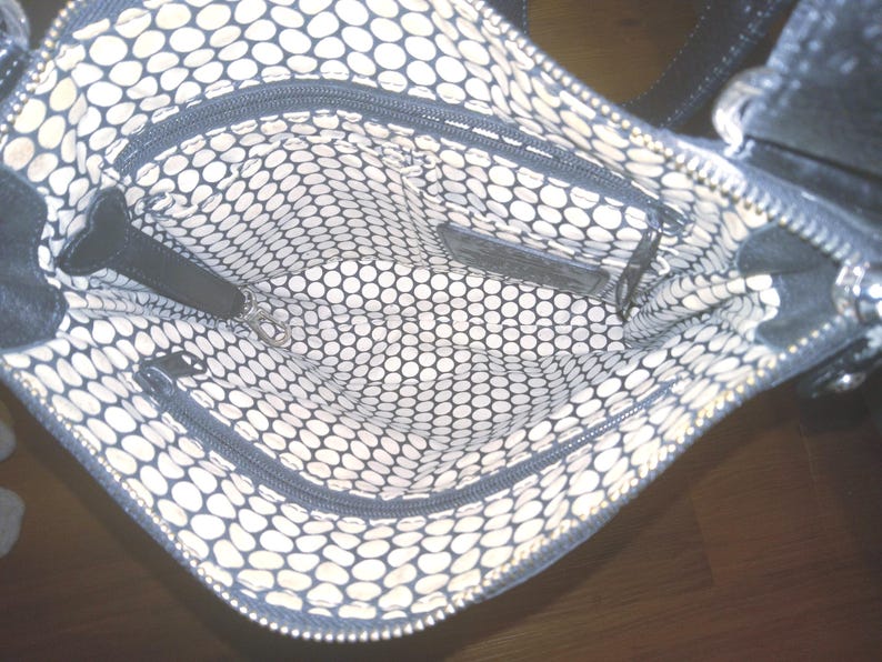 May include: Black handbag with a patterned interior. The interior features a white and black polka dot design. The bag has multiple zippered compartments and a black leather strap. The bag is open, revealing the interior.
