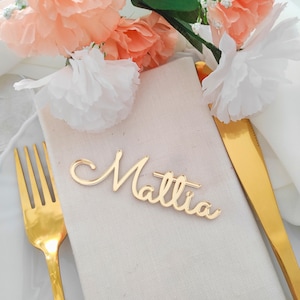 Può includere: Un segnaposto dorato con il nome "Mattia" in corsivo è appoggiato su un tovagliolo beige piegato. Una forchetta e un coltello dorati sono su entrambi i lati. Fiori color pesca e bianchi sullo sfondo.