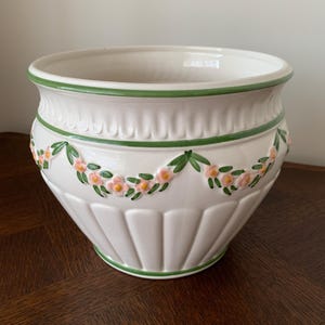 May include: A white ceramic planter featuring a scalloped rim and a base with vertical fluting. The exterior is decorated with a raised relief garland of small pink flowers and green leaves, accented by thin green painted trim lines.