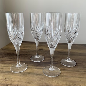 May include: Four clear crystal champagne flutes with detailed cut glass designs. The flutes feature a tall, slim shape with a diamond pattern near the base and vertical cuts above. They are displayed on a wooden surface.