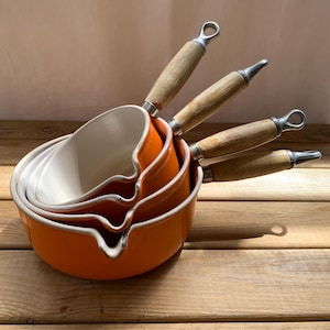 May include: A set of four nested orange ceramic saucepans with cream-coloured interiors and light wooden handles. Each pan features a distinct pouring spout on the rim and a metal loop at the end of the handle for hanging, arranged in a stack.