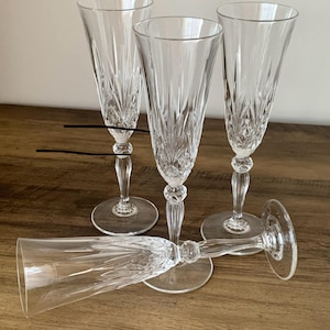 May include: Four clear glass champagne flutes featuring a vertical cut pattern on the bowl and a multi-faceted stem. Three of the flutes are standing upright, while one flute is positioned horizontally in the foreground.