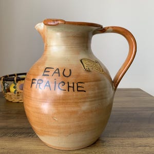 May include: A ceramic jug in shades of brown and beige, with the words "EAU FRAICHE" painted on the front. It features a handle and a spout. A small label is attached to the side. The jug is likely for serving water.