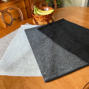 May include: Three table runners in different shades of gray, from light to dark, laid out on a wooden table. The runners are made of a woven fabric with a subtle texture.