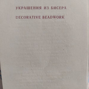May include: A book cover with the title "УКРАШЕНИЯ ИЗ БИСЕРА" in maroon letters above "DECORATIVE BEADWORK". The background is a light beige color. The book appears to be about decorative beadwork.