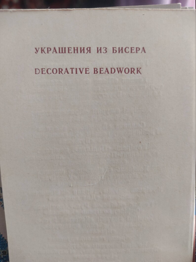 May include: A book cover with the title "УКРАШЕНИЯ ИЗ БИСЕРА" in maroon letters above "DECORATIVE BEADWORK". The background is a light beige color. The book appears to be about decorative beadwork.