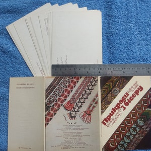 May include: A collection of vintage books and postcards on a blue background. The books feature decorative beadwork designs with text in a foreign language. A ruler is placed next to the items, and a stack of postcards is visible.