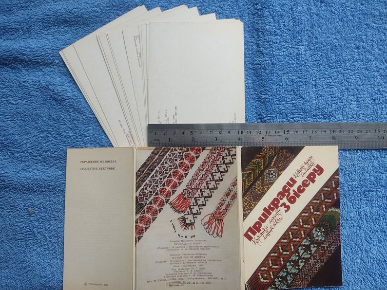 May include: A collection of vintage books and postcards on a blue background. The books feature decorative beadwork designs with text in a foreign language. A ruler is placed next to the items, and a stack of postcards is visible.