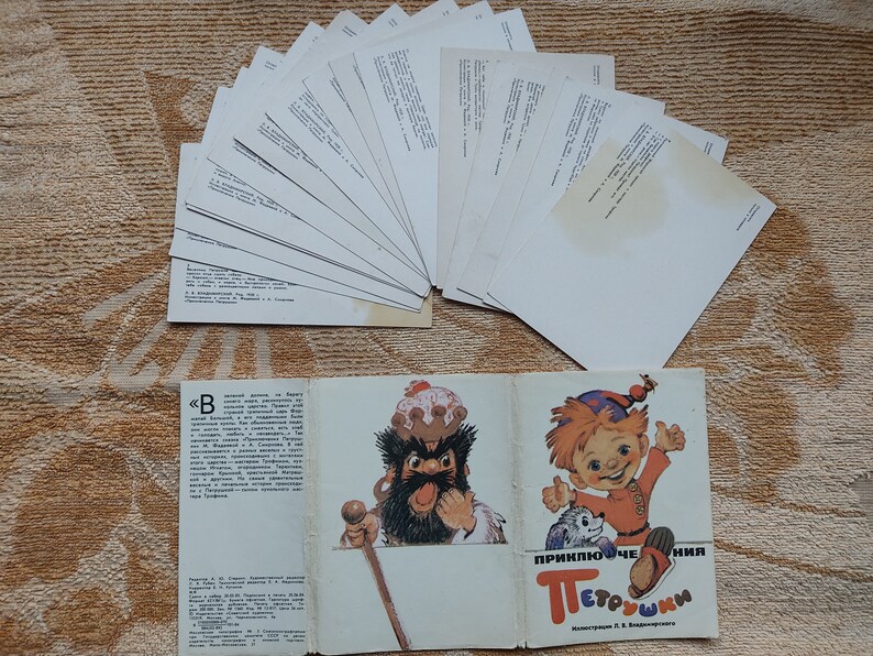 May include: A vintage book titled "Приключения Петрушки" (Adventures of Petrushka) with illustrations of a bearded man in a crown and a red-haired boy with a dog. The book is open to reveal the text and illustrations. There are also 15 blank postcards in the background.