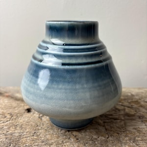 May include: A small, blue ceramic vase with a rounded body and a narrow neck. The vase has a gradient effect, transitioning from a dark blue at the top to a lighter blue at the base. The vase is approximately 5 inches tall.