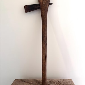 May include: A rustic hand tool featuring a long, weathered wooden handle that splits into a V-shape at the top. A dark, oxidised metal blade is inserted horizontally through the wood, extending from one side of the handle head.