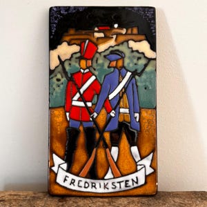 May include: A rectangular ceramic wall plaque featuring a colourful enamel illustration of two soldiers in historical uniforms, one in red and one in blue, standing before a fortress. A white banner at the bottom displays the text FREDRIKSTEN.