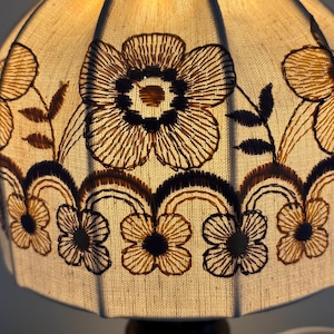 Pode incluir: Um abajur de tecido na cor creme com padr&otilde;es florais bordados em castanho escuro. O design inclui flores centrais grandes e uma borda inferior de flores menores conectadas por linhas arqueadas, iluminado por dentro por uma luz quente.