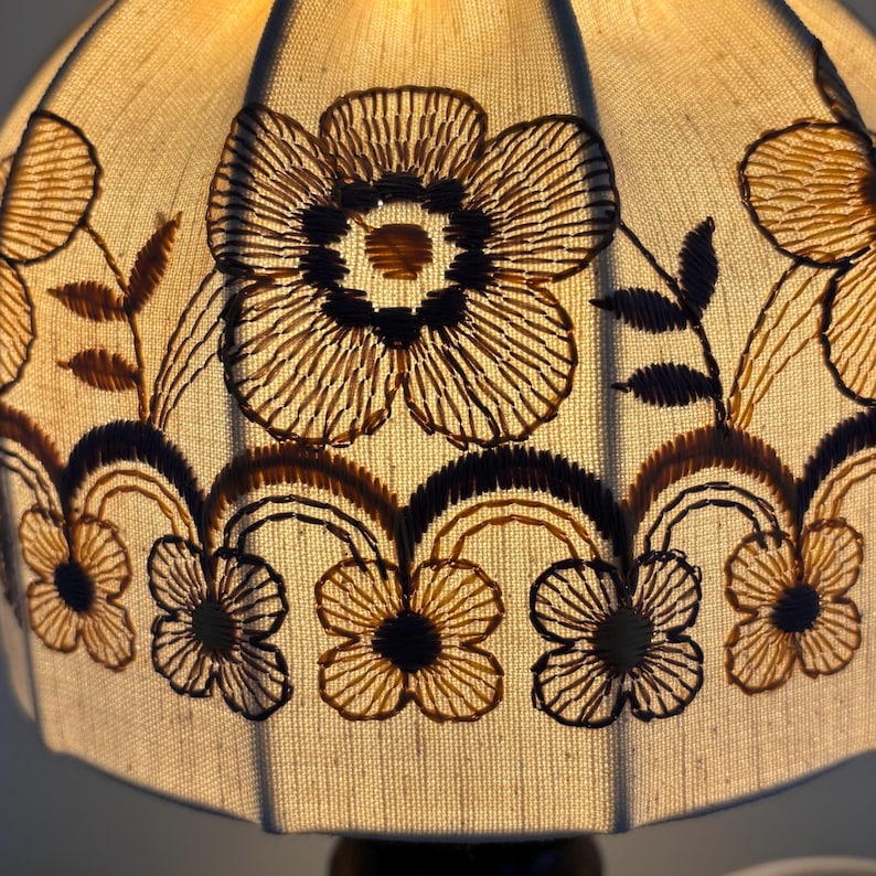 Pode incluir: Um abajur de tecido na cor creme com padr&otilde;es florais bordados em castanho escuro. O design inclui flores centrais grandes e uma borda inferior de flores menores conectadas por linhas arqueadas, iluminado por dentro por uma luz quente.