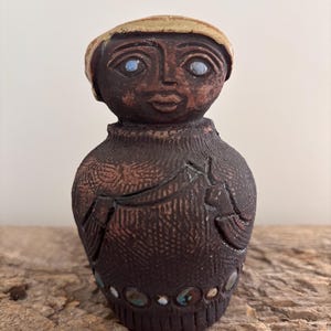 Może przedstawiać: Ręcznie wykonana ceramiczna figurka przedstawiająca osobę o ciemnobrązowej karnacji, niebieskich oczach i beżowym nakryciu głowy. Figurka ma teksturowany, ciemnobrązowy korpus z dekoracyjnymi akcentami i rzędem kolorowych ozdób u podstawy. Figurka jest eksponowana na drewnianej powierzchni.