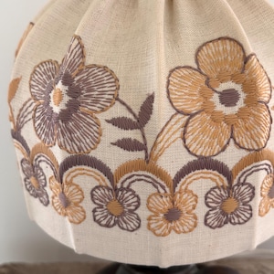 Pode incluir: Um abajur de tecido na cor creme com um padr&atilde;o floral bordado. O design inclui flores grandes em tons de castanho e bege, acentuadas por motivos florais menores e repetitivos ao longo da borda inferior em um arranjo festonado.