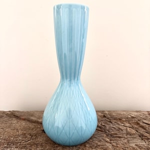Puede incluir: Un jarrón de cerámica azul claro con una base bulbosa y un cuello delgado. El jarrón tiene una superficie texturizada con líneas verticales en el cuello y un patrón de diamantes en la base. El jarrón mide unos 25 cm de alto.