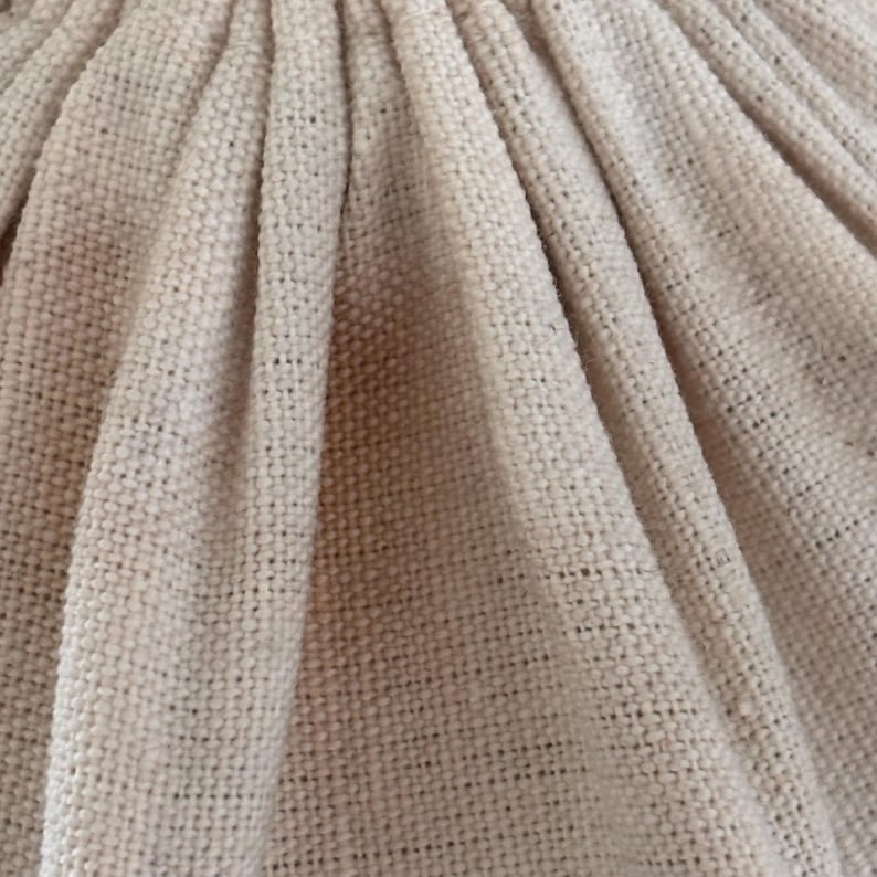 Pode incluir: Um close-up de tecido bege claro com uma textura tecida vis&iacute;vel. O material &eacute; reunido em dobras verticais, criando uma s&eacute;rie de vincos profundos e r&iacute;tmicos que correm de cima para baixo, destacando a trama do tecido.