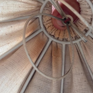 Pode incluir: Um interior de c&uacute;pula de l&acirc;mpada em tecido bege com uma estrutura de arame met&aacute;lico branco. O tecido &eacute; franzido no centro superior em torno de um anel met&aacute;lico circular. O material apresenta descolora&ccedil;&otilde;es e manchas castanho-claras vis&iacute;veis.