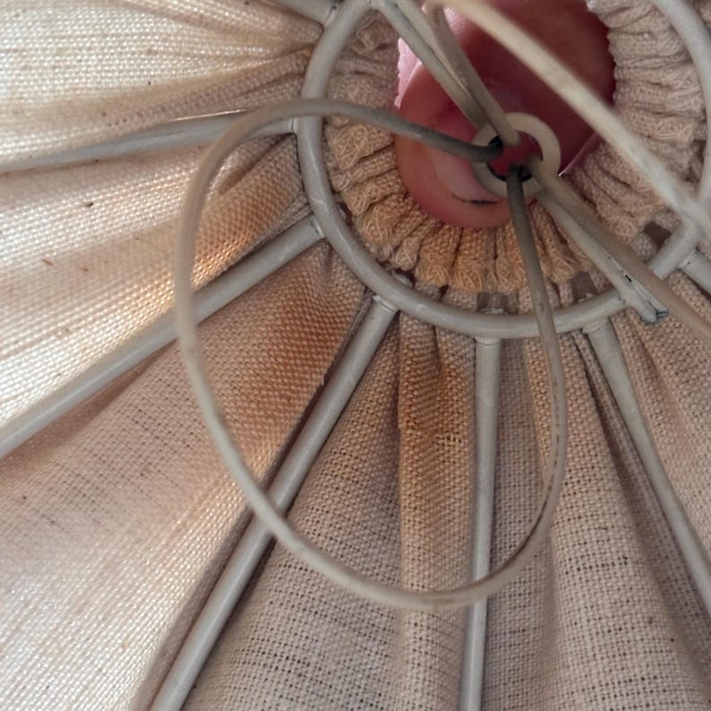 Pode incluir: Um interior de c&uacute;pula de l&acirc;mpada em tecido bege com uma estrutura de arame met&aacute;lico branco. O tecido &eacute; franzido no centro superior em torno de um anel met&aacute;lico circular. O material apresenta descolora&ccedil;&otilde;es e manchas castanho-claras vis&iacute;veis.