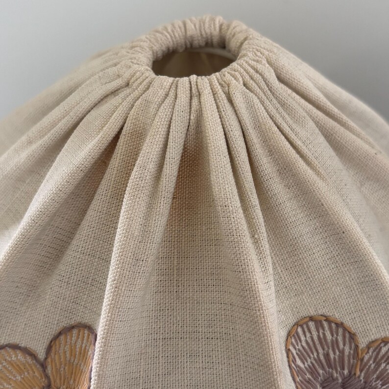 Pode incluir: Um abajur de tecido bege com uma abertura superior franzida e el&aacute;stica. O material tecido e texturizado apresenta motivos florais bordados em tons de amarelo suave e roxo delicado, criando uma apar&ecirc;ncia plissada e estruturada ao redor do topo.