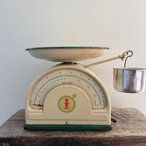 Op de afbeelding: Vintage crèmekleurige keukenweegschaal met groene rand en een ronde wijzerplaat. De weegschaal heeft een metalen kom en een kleine hangende beker. De wijzerplaat geeft tot 10 kg aan. Met groen en rood embleem.