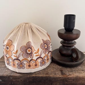 Pode incluir: Um abajur de tecido bege com padr&otilde;es florais bordados em castanho e laranja e topo franzido, ao lado de uma base de candeeiro de madeira castanho-escura com casquilho preto, sobre uma superf&iacute;cie de madeira texturizada.