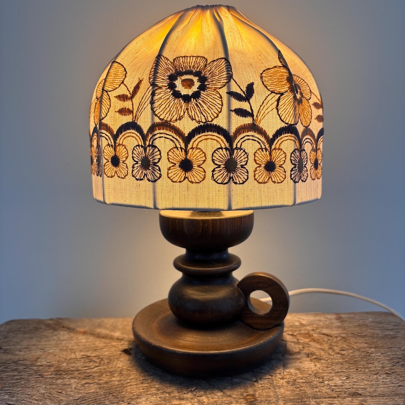 Pode incluir: Um candeeiro de mesa com base de madeira castanho-escura com uma pega circular e um abajur de tecido em forma de c&uacute;pula. O abajur em tom cru &eacute; decorado com padr&otilde;es florais bordados a castanho e uma borda recortada, emitindo um brilho quente.