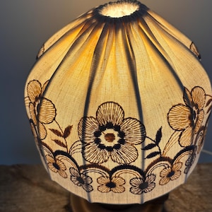 Pode incluir: Um abajur de tecido em forma de c&uacute;pula com um design plissado e franzido, que emite uma luz quente e brilhante. O material branco sujo &eacute; decorado com padr&otilde;es florais bordados em castanho escuro e motivos de vinhas entrela&ccedil;adas.