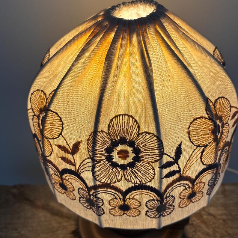 Pode incluir: Um abajur de tecido em forma de c&uacute;pula com um design plissado e franzido, que emite uma luz quente e brilhante. O material branco sujo &eacute; decorado com padr&otilde;es florais bordados em castanho escuro e motivos de vinhas entrela&ccedil;adas.