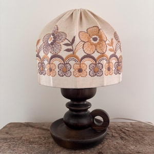 Pode incluir: Um candeeiro de mesa com base de madeira castanho-escura com uma pega circular e um abajur de tecido em forma de c&uacute;pula. O abajur bege &eacute; decorado com padr&otilde;es florais bordados em tons de laranja, castanho e bronze, com uma borda festonada.