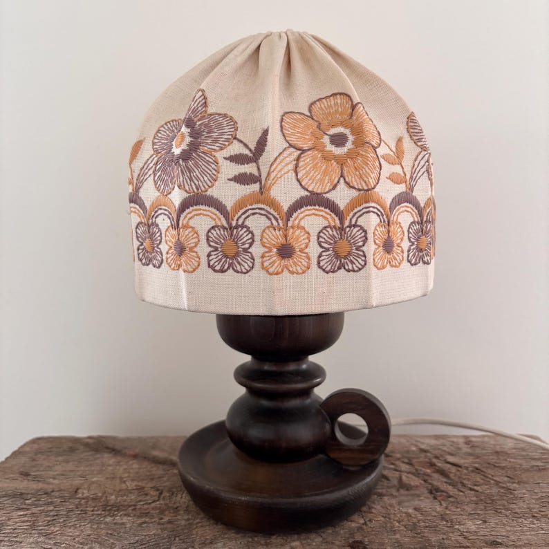 Pode incluir: Um candeeiro de mesa com base de madeira castanho-escura com uma pega circular e um abajur de tecido em forma de c&uacute;pula. O abajur bege &eacute; decorado com padr&otilde;es florais bordados em tons de laranja, castanho e bronze, com uma borda festonada.