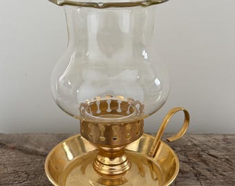 Puede incluir: Un candelabro de latón vintage con una pantalla de vidrio transparente. El candelabro tiene una base redonda, un tallo decorativo y un asa. La pantalla de vidrio tiene un borde festoneado.