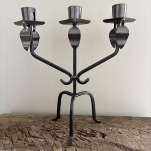 May include: A dark grey metal candelabra featuring three candle holders arranged in a row. Each holder is supported by a curved arm adorned with a small metal leaf detail. The structure stands on a tripod base made of three curved metal legs.