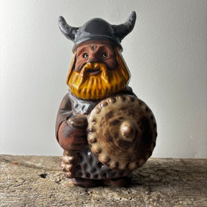 May include: A ceramic figurine of a Viking warrior with a brown beard and a horned helmet. The warrior is holding a shield with a brown and white design.