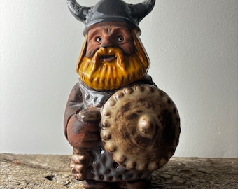 May include: A ceramic figurine of a Viking warrior with a brown beard and a horned helmet. The warrior is holding a shield with a brown and white design.