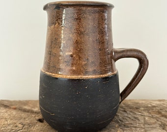 May include: A brown and black ceramic pitcher with a handle. The pitcher has a speckled glaze and a black base.