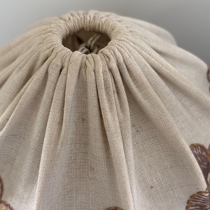 Pode incluir: Uma bolsa com cord&atilde;o feita de tecido bege, apresentando um fecho franzido na parte superior com um cord&atilde;o a condizer. O exterior da bolsa &eacute; decorado com um padr&atilde;o repetitivo de motivos circulares castanhos texturizados ao longo da borda inferior.