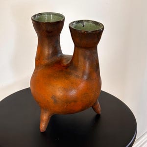 Könnte beinhalten: Ein Keramikgefäß mit zwei Ausgüssen, einem abgerundeten Boden und drei kurzen Beinen. Die Außenseite hat ein fleckiges, erdiges Braun- und Orange-Finish, während das Innere jedes Ausgusses mit einer kontrastierenden, hellen, mintgrünen Glasur versehen ist.