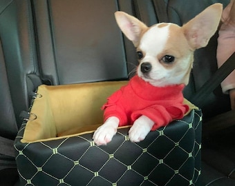 May include: A small chihuahua dog wearing a red sweater sits in a black and gold pet car seat. The seat has a diamond pattern and is secured with straps.